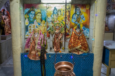 A temple-like setting featuring multiple statues adorned with colorful fabrics and garlands. Behind them are vibrant paintings depicting deities, with one prominent figure holding a trident. A hanging metal object, possibly a lamp or incense holder, is positioned in front of the statues.