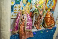 Hindu deities are adorned in intricate, colorful fabrics with golden embellishments, surrounded by garlands and decorative items, displayed against a backdrop of vibrant religious posters.