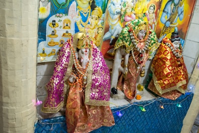 Hindu deities are adorned in intricate, colorful fabrics with golden embellishments, surrounded by garlands and decorative items, displayed against a backdrop of vibrant religious posters.