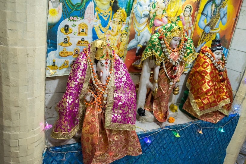 Hindu deities are adorned in intricate, colorful fabrics with golden embellishments, surrounded by garlands and decorative items, displayed against a backdrop of vibrant religious posters.