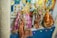 Hindu deities are adorned in intricate, colorful fabrics with golden embellishments, surrounded by garlands and decorative items, displayed against a backdrop of vibrant religious posters.
