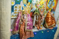 Hindu deities are adorned in intricate, colorful fabrics with golden embellishments, surrounded by garlands and decorative items, displayed against a backdrop of vibrant religious posters.