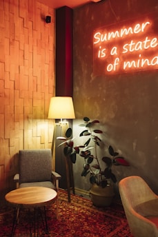 A cozy interior space featuring a warm wooden wall, a grey upholstered chair, a small round wooden table, a potted plant, and a floor lamp. A neon sign on the wall reads 'Summer is a state of mind' in glowing orange light, adding an ambient mood to the room. The floor is covered with a richly patterned red rug.