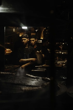 Two male chefs wearing black uniforms and caps are working in a dimly lit kitchen. One of them is holding a paper bag and appears to be focused on his task while the other watches. The background shows various kitchen equipment and utensils.