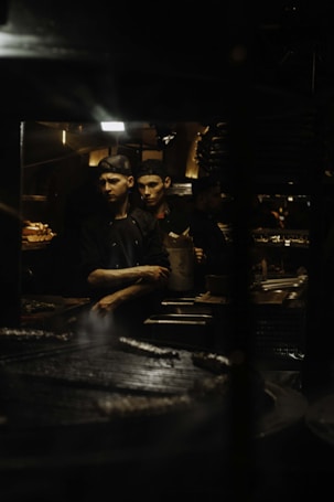 Two male chefs wearing black uniforms and caps are working in a dimly lit kitchen. One of them is holding a paper bag and appears to be focused on his task while the other watches. The background shows various kitchen equipment and utensils.