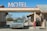 A vintage car is parked under the awning of an old-fashioned motel, with a large sign reading 'MOTEL' on top. The structure features stone walls and a small payphone is visible on the right side. The setting is in a desert landscape, with dry bushes and a clear blue sky in the background.
