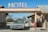 A vintage car is parked under the awning of an old-fashioned motel, with a large sign reading 'MOTEL' on top. The structure features stone walls and a small payphone is visible on the right side. The setting is in a desert landscape, with dry bushes and a clear blue sky in the background.