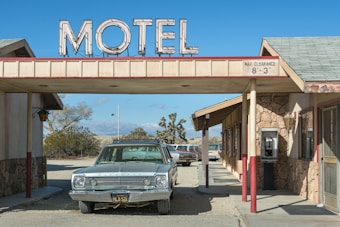 A vintage car is parked under the awning of an old-fashioned motel, with a large sign reading 'MOTEL' on top. The structure features stone walls and a small payphone is visible on the right side. The setting is in a desert landscape, with dry bushes and a clear blue sky in the background.