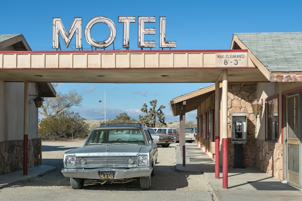 A vintage car is parked under the awning of an old-fashioned motel, with a large sign reading 'MOTEL' on top. The structure features stone walls and a small payphone is visible on the right side. The setting is in a desert landscape, with dry bushes and a clear blue sky in the background.