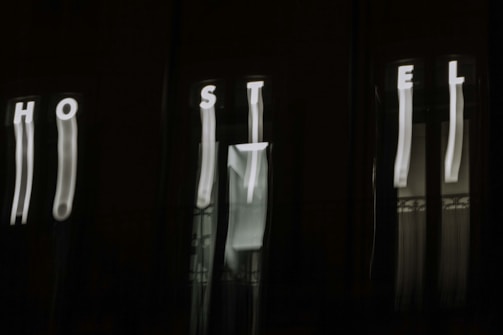 The image features a dark background with blurry, vertically elongated white letters spelling out 'HOSTEL'. The letters appear to be on an illuminated sign, but due to the blur, they are distorted and stretched.