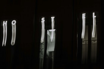 The image features a dark background with blurry, vertically elongated white letters spelling out 'HOSTEL'. The letters appear to be on an illuminated sign, but due to the blur, they are distorted and stretched.