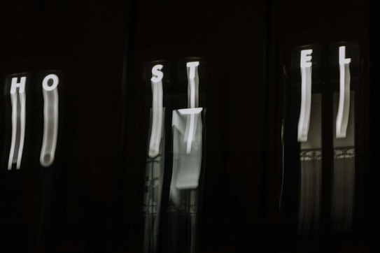 The image features a dark background with blurry, vertically elongated white letters spelling out 'HOSTEL'. The letters appear to be on an illuminated sign, but due to the blur, they are distorted and stretched.