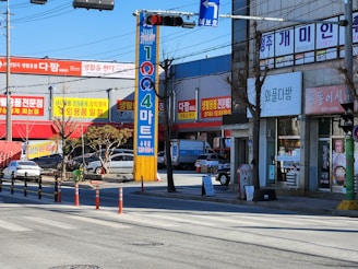 An urban street scene featuring colorful signage on buildings advertising various businesses, such as a mart and other shops. The image shows a crosswalk with traffic lights and signage written in Korean. There are cars parked on the side of the street and some bare trees, indicating an urban commercial area.