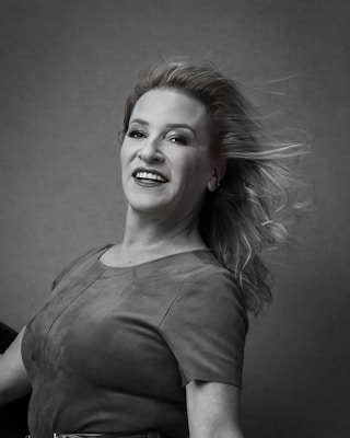 A monochrome portrait of a smiling woman with flowing hair, wearing a short-sleeved dress. The background is plain and neutral, highlighting her expression and dynamic pose.