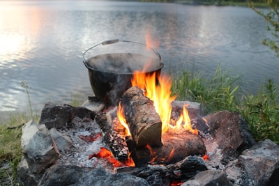 A campfire is burning by a lakeside, with flames licking the edges of a large pot suspended over the fire. The fire is surrounded by a ring of stones, with ash and embers visible. The serene lake in the background reflects the soft light of the sky.