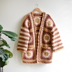 A crocheted cardigan with a geometric pattern in shades of brown, yellow, and cream hangs neatly on a curved wooden hanger. The design features circular motifs framed by stripes, giving a retro and handcrafted appearance. In the background, a green leafy plant adds a touch of natural contrast against a light-colored wall.