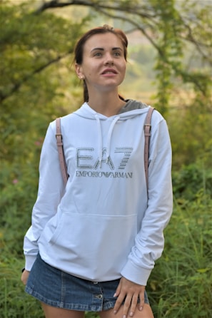 A person is standing outdoors in a lush green environment. They are wearing a white hoodie with the text 'Emporio Armani' and a denim skirt. The background is filled with greenery and blurred foliage.