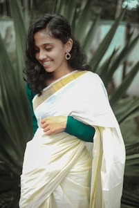 A woman in a traditional white and gold saree is smiling, with surrounding lush green foliage in the background. Her hair is styled in loose waves, and she is wearing ornate earrings.