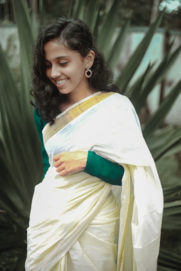 A woman in a traditional white and gold saree is smiling, with surrounding lush green foliage in the background. Her hair is styled in loose waves, and she is wearing ornate earrings.