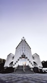 A modern church building with a striking architectural design featuring vertical lines and a large cross at the peak. The structure is symmetrical, with two wings on either side and a staircase leading to the entrance. The sky above is clear with a gradient from light to dark.