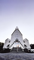 A modern church building with a striking architectural design featuring vertical lines and a large cross at the peak. The structure is symmetrical, with two wings on either side and a staircase leading to the entrance. The sky above is clear with a gradient from light to dark.