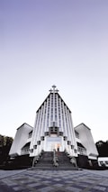 A modern church building with a striking architectural design featuring vertical lines and a large cross at the peak. The structure is symmetrical, with two wings on either side and a staircase leading to the entrance. The sky above is clear with a gradient from light to dark.