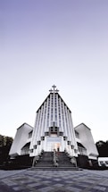 A modern church building with a striking architectural design featuring vertical lines and a large cross at the peak. The structure is symmetrical, with two wings on either side and a staircase leading to the entrance. The sky above is clear with a gradient from light to dark.