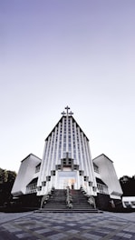 A modern church building with a striking architectural design featuring vertical lines and a large cross at the peak. The structure is symmetrical, with two wings on either side and a staircase leading to the entrance. The sky above is clear with a gradient from light to dark.