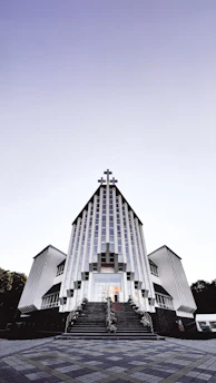A modern church building with a striking architectural design featuring vertical lines and a large cross at the peak. The structure is symmetrical, with two wings on either side and a staircase leading to the entrance. The sky above is clear with a gradient from light to dark.