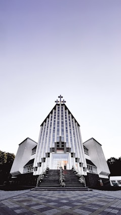 A modern church building with a striking architectural design featuring vertical lines and a large cross at the peak. The structure is symmetrical, with two wings on either side and a staircase leading to the entrance. The sky above is clear with a gradient from light to dark.