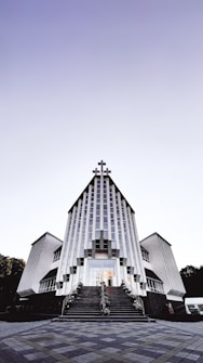 A modern church building with a striking architectural design featuring vertical lines and a large cross at the peak. The structure is symmetrical, with two wings on either side and a staircase leading to the entrance. The sky above is clear with a gradient from light to dark.
