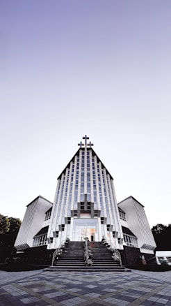 A modern church building with a striking architectural design featuring vertical lines and a large cross at the peak. The structure is symmetrical, with two wings on either side and a staircase leading to the entrance. The sky above is clear with a gradient from light to dark.