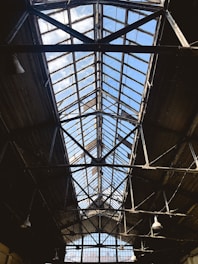 A large industrial ceiling made of metal and glass, with a triangular structure leading upwards toward a bright blue sky. The framework is dark, contrasting with the sunlight filtering through the glass panels. The ceiling displays signs of age and wear, with rustic elements and a few visible light fixtures hanging down.