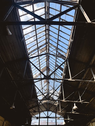 A large industrial ceiling made of metal and glass, with a triangular structure leading upwards toward a bright blue sky. The framework is dark, contrasting with the sunlight filtering through the glass panels. The ceiling displays signs of age and wear, with rustic elements and a few visible light fixtures hanging down.