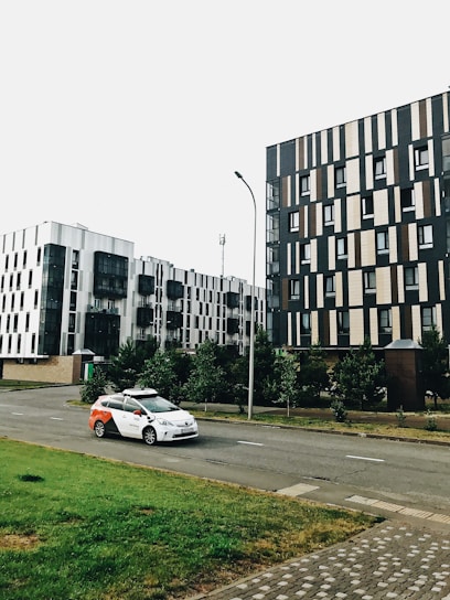 A modern urban scene featuring two contemporary apartment buildings with geometrically patterned facades. A white and red taxi is parked on the road. The setting includes a sidewalk and a small grassy area with young trees lining the street. The overcast sky creates a muted lighting.