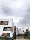 A modern residential building with a predominantly white facade accented by wooden panels. The architecture features clean lines and multiple windows, contributing to a contemporary design. The building is surrounded by small trees and shrubs, and the sky above is cloudy, suggesting overcast weather.