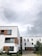 A modern residential building with a predominantly white facade accented by wooden panels. The architecture features clean lines and multiple windows, contributing to a contemporary design. The building is surrounded by small trees and shrubs, and the sky above is cloudy, suggesting overcast weather.