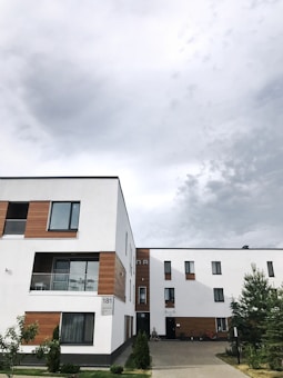 A modern residential building with a predominantly white facade accented by wooden panels. The architecture features clean lines and multiple windows, contributing to a contemporary design. The building is surrounded by small trees and shrubs, and the sky above is cloudy, suggesting overcast weather.
