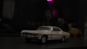 A model car is placed in low light with a dark, blurred background. The car is cream-colored and appears to have a classic design, possibly from the 1960s or 1970s.