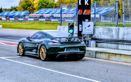 A sleek sports car with golden wheels accelerates on a racing track. The vehicle is captured from behind, displaying a glossy, dark exterior. The track features vibrant blue seating in the background, surrounded by green foliage. A traffic light shows green, indicating the car is allowed to proceed.
