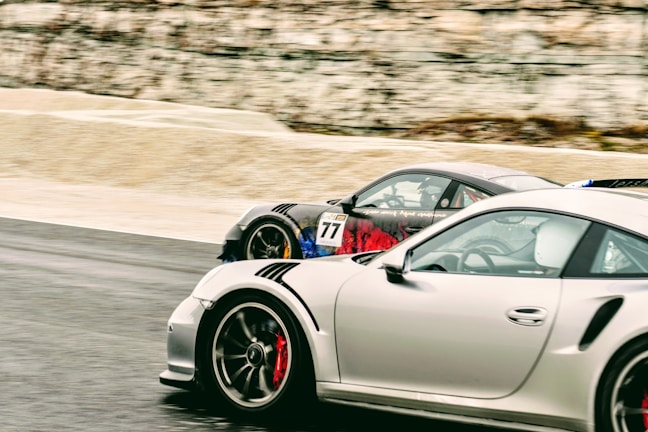 Two sports cars in vibrant colors speed along a racetrack. The cars are closely aligned, indicating an intense race. Distinctive decals and aerodynamic features are visible, highlighting the performance-oriented design. The background is slightly blurred, emphasizing the motion and speed of the vehicles.