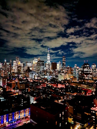 A vibrant city skyline at night with numerous illuminated skyscrapers under a partly cloudy sky. The city lights create a vivid and bustling atmosphere.