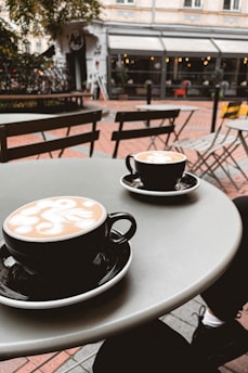 Two black coffee cups with intricate latte art sit on a rounded table in an outdoor café setting. In the background, there is a European-style building with large windows and some greenery. The atmosphere is relaxed and inviting, with a few empty chairs and tables nearby.
