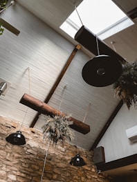 A rustic interior with a wooden ceiling and decorative hanging plants. The ceiling features beams in a diagonal design, interspersed with hanging light fixtures. Stone walls add to the rustic charm, while hanging plants provide a natural touch.