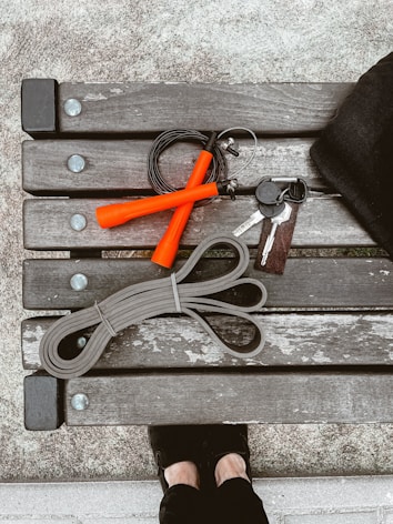 A wooden bench with metal bolts is viewed from above, holding fitness-related items including a jump rope with orange handles, an elastic exercise band, and a keychain with keys. The pavement and a pair of feet in black shoes are partially visible.