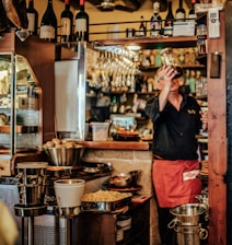 A cozy bar setting features a bartender in a red apron holding up a glass jar. The shelves are stocked with various bottles of wine and jars, while glasses hang overhead. The counter is cluttered with kitchen utensils, bowls, and buckets.