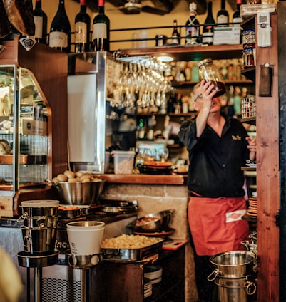 A cozy bar setting features a bartender in a red apron holding up a glass jar. The shelves are stocked with various bottles of wine and jars, while glasses hang overhead. The counter is cluttered with kitchen utensils, bowls, and buckets.