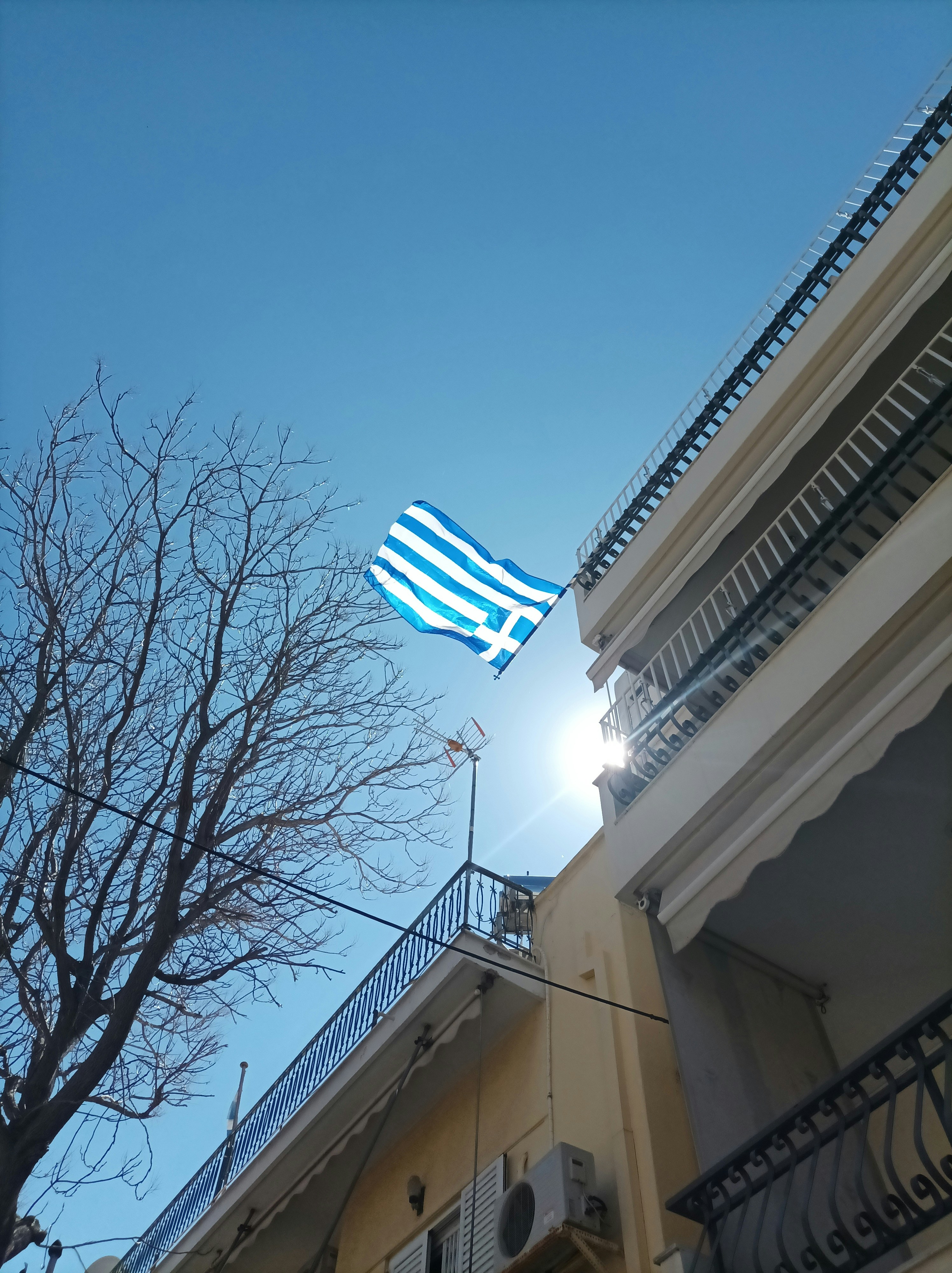 A Greek flag flutters against a clear blue sky, with the sun peeking over the top of a building. The shot is taken from a low angle, showing a bare tree and the detailed balconies of the adjacent buildings.