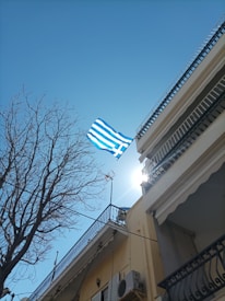 A Greek flag flutters against a clear blue sky, with the sun peeking over the top of a building. The shot is taken from a low angle, showing a bare tree and the detailed balconies of the adjacent buildings.