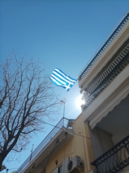A Greek flag flutters against a clear blue sky, with the sun peeking over the top of a building. The shot is taken from a low angle, showing a bare tree and the detailed balconies of the adjacent buildings.
